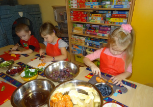 Troje dzieci siedzi przy stole, w ręku trzymają noże, którymi kroją owoce na desce. Na środku stołu stoją miski wypełnione owocami.